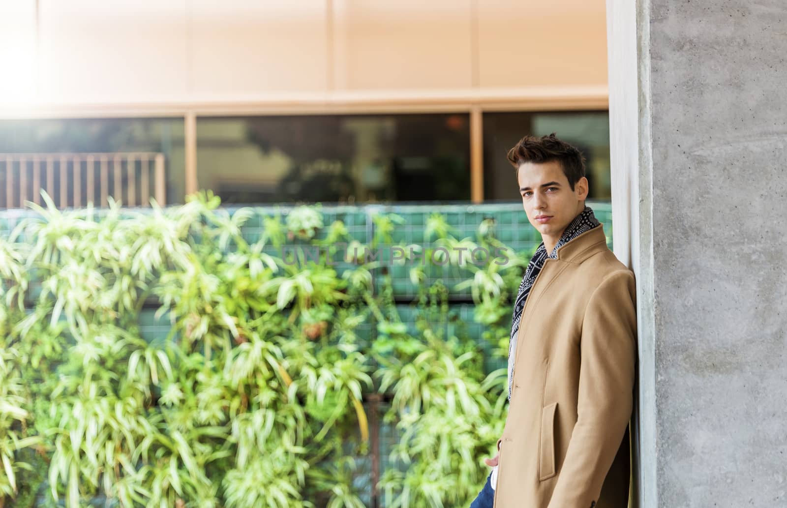 Side view of young trendy man wearing denim clothes leaning on a wall while looking camera outdoors in sunny day