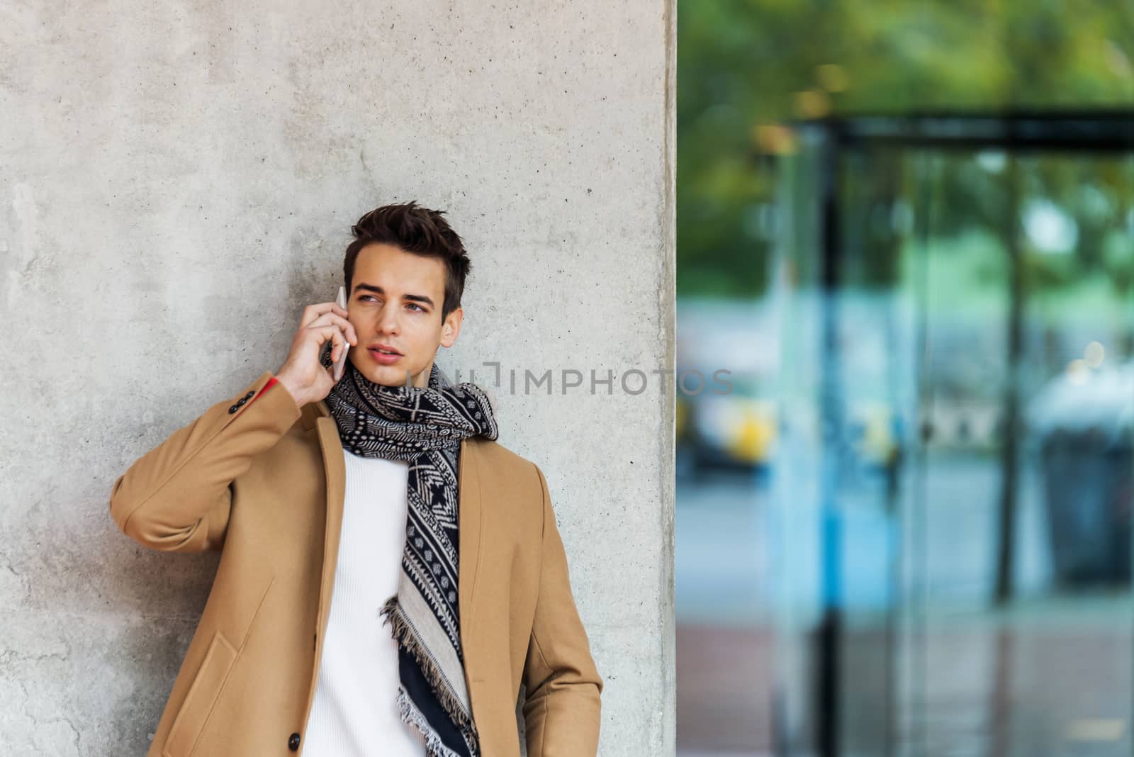 Front view of stylish young man wearing denim clothes leaning on a wall while using a mobile phone outdoors by raferto1973