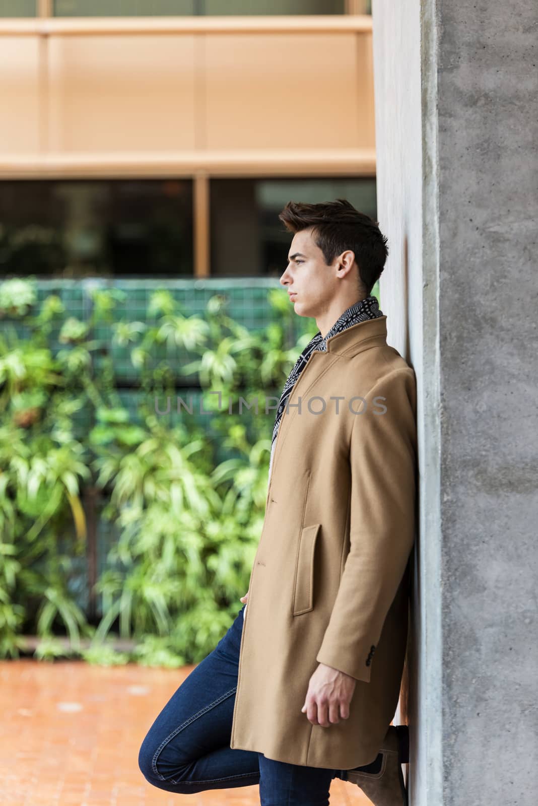 Side view of stylish young man wearing coat and scarf leaning on a wall while looking away