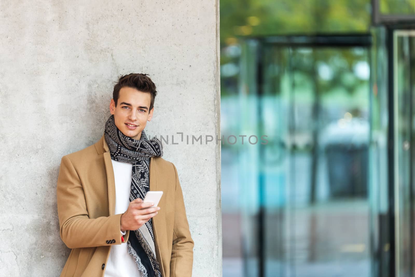 Front view of stylish young man wearing denim clothes leaning on a wall while using a mobile phone outdoors by raferto1973