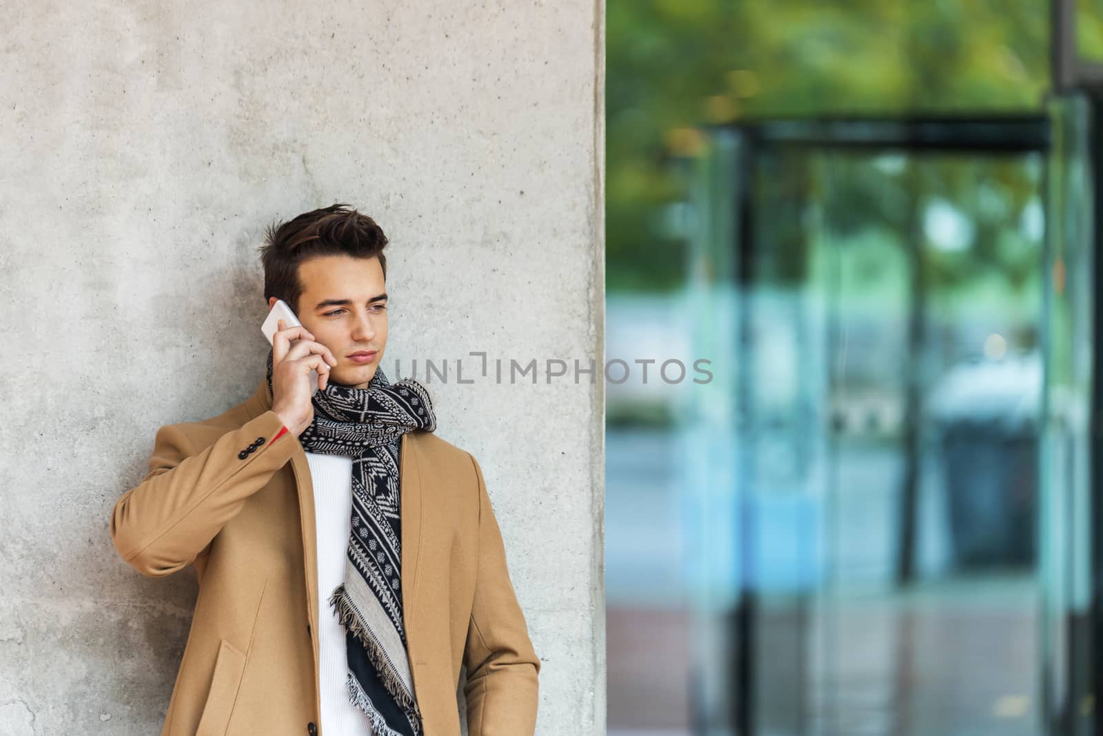 Front view of stylish young man wearing denim clothes leaning on a wall while using a mobile phone outdoors by raferto1973