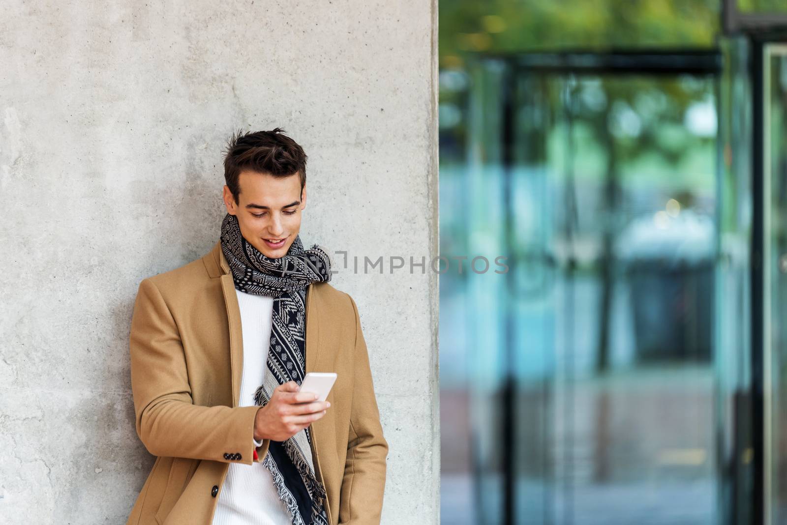 Front view of stylish young man wearing denim clothes leaning on a wall while using a mobile phone outdoors by raferto1973