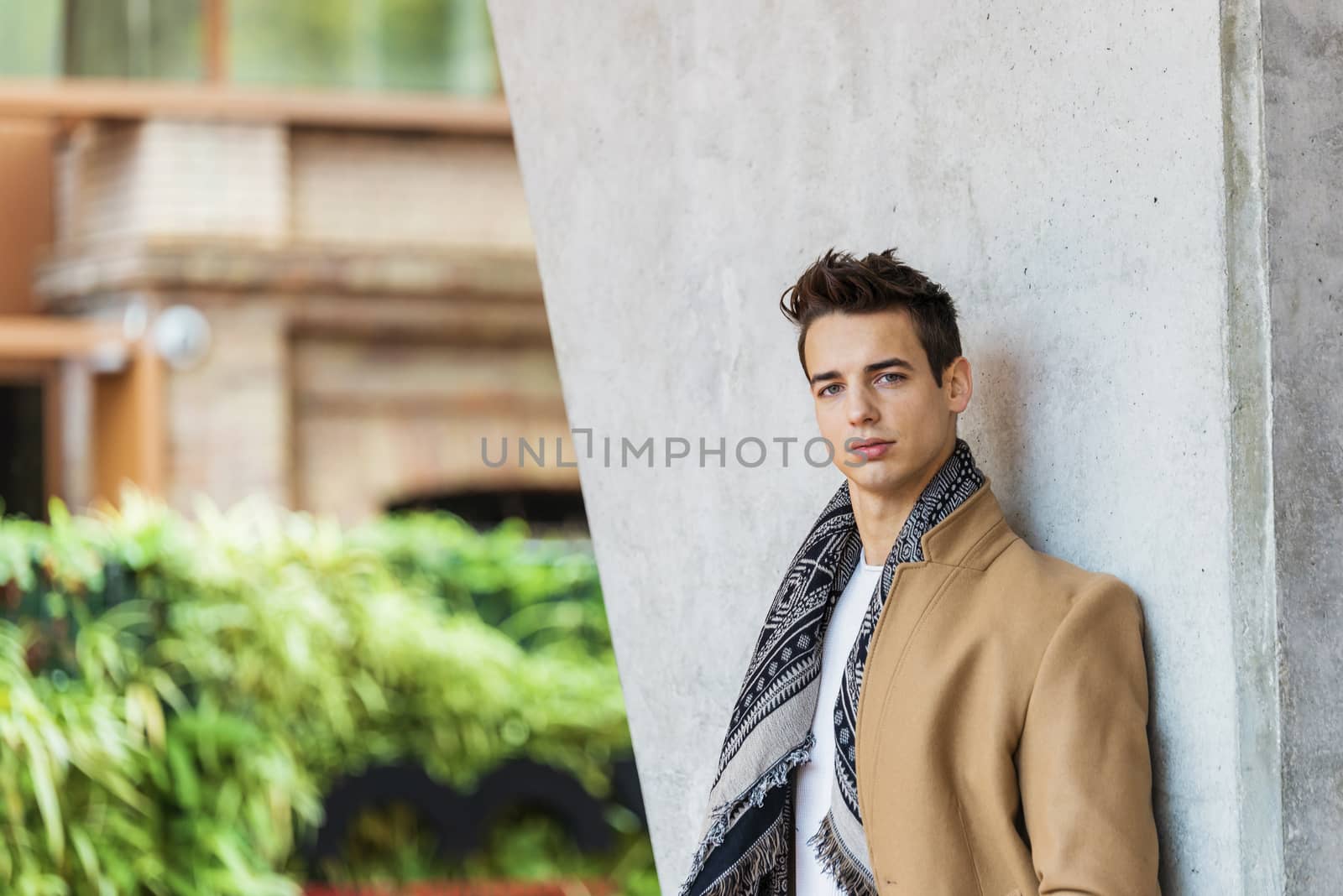 Side view of young trendy man wearing denim clothes leaning on a wall while looking camera outdoors in sunny day