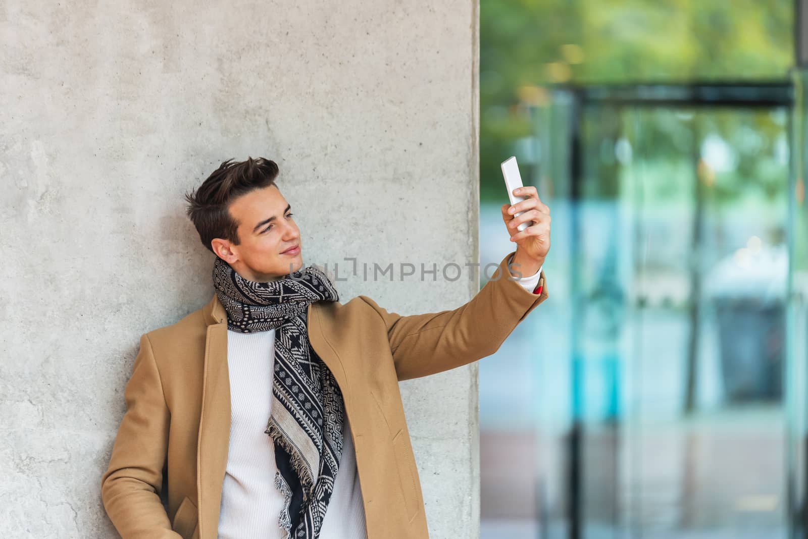 Side view of young trendy man leaning on a wall outdoors wearing denim clothes while taking a selfie in sunny day