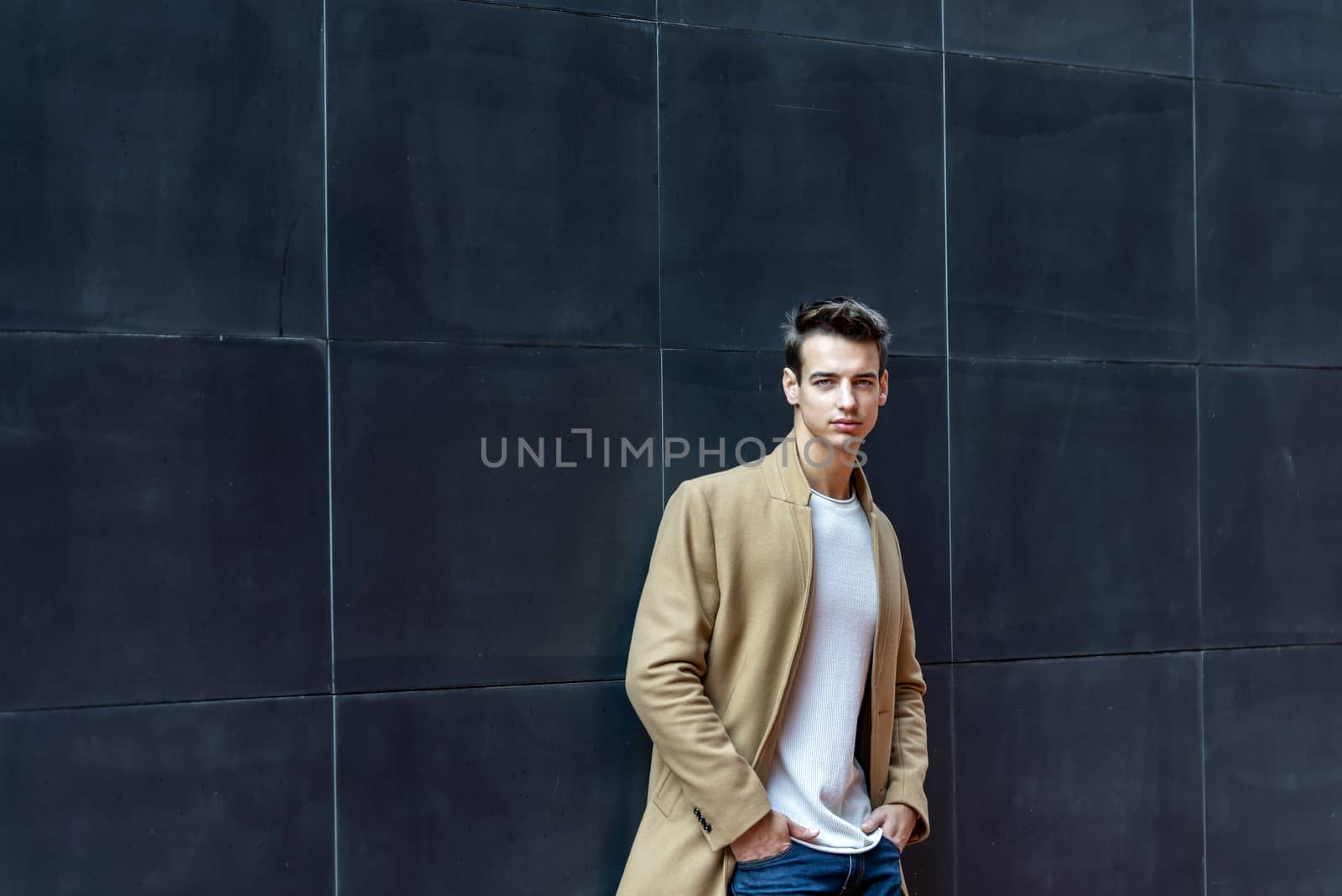 Front view of a trendy young man standing against black wall while looking camera outdoors in the street