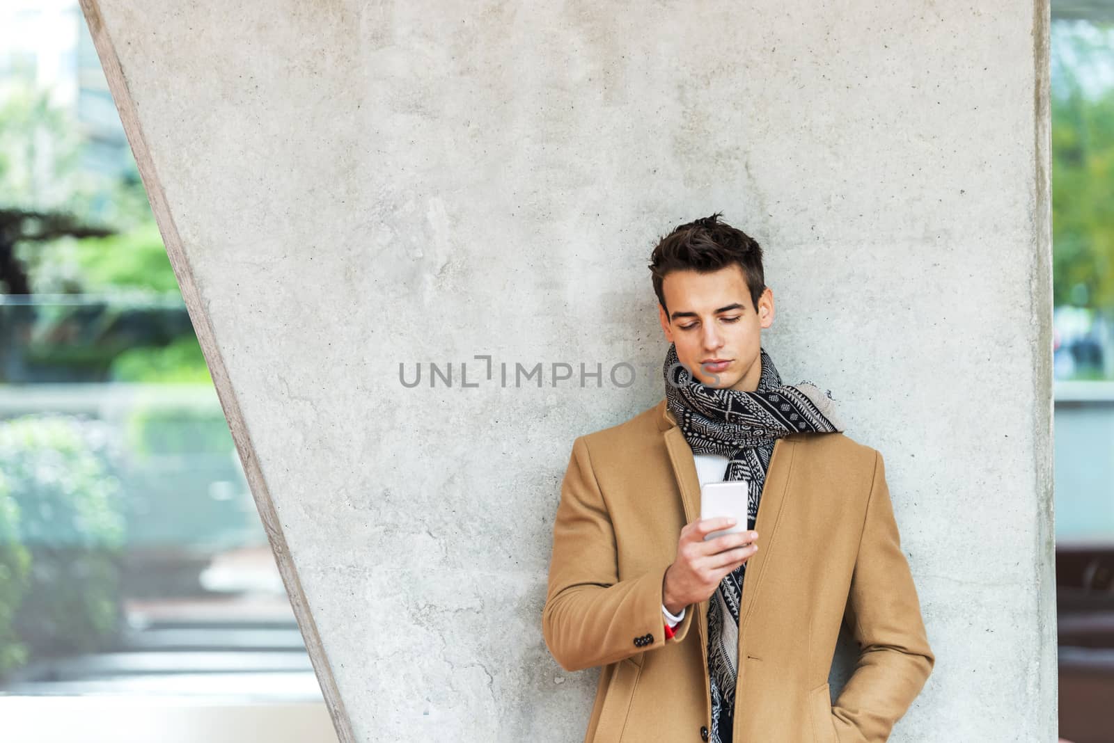 Front view of stylish young man wearing denim clothes leaning on a wall while using a mobile phone outdoors by raferto1973