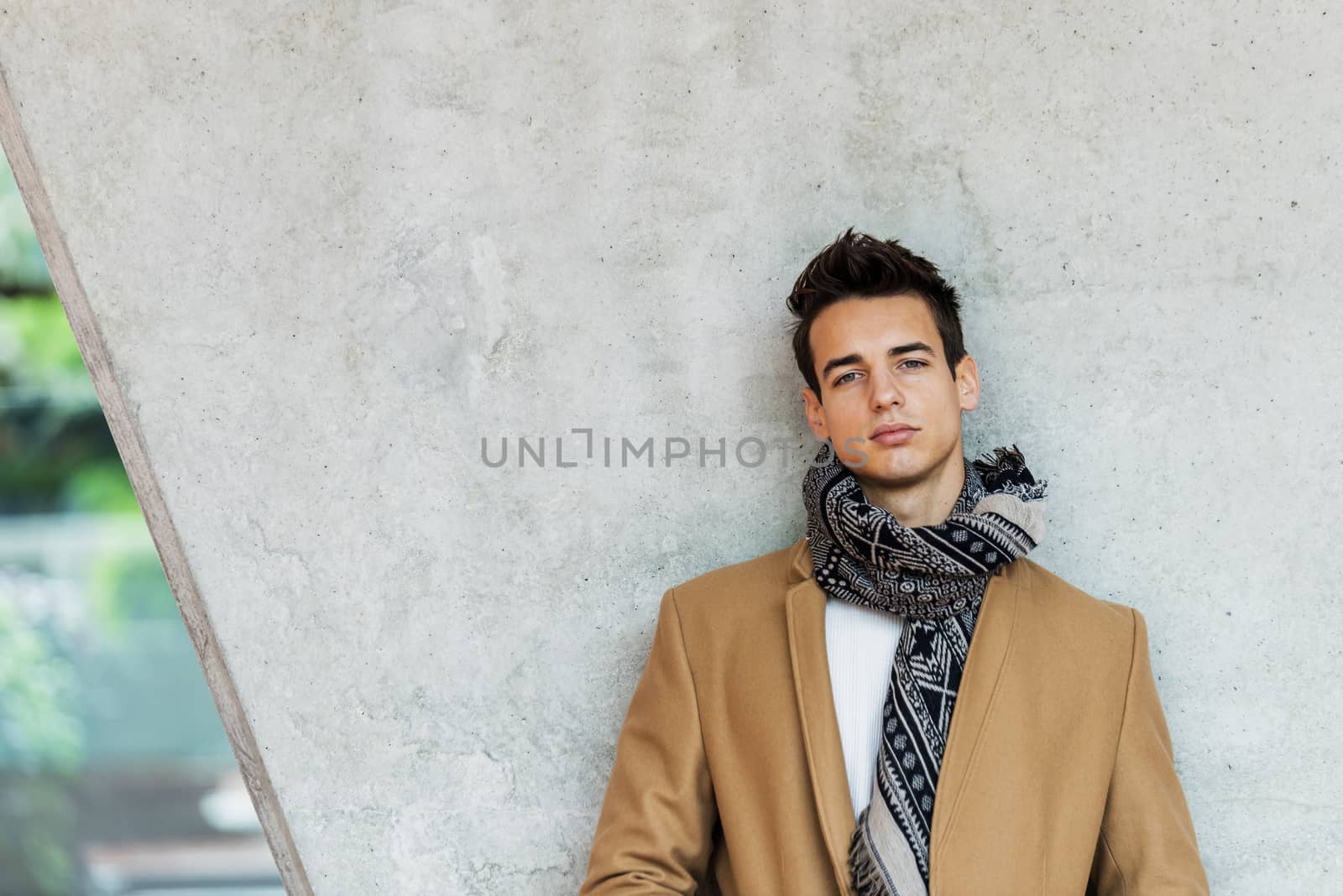 Front view of stylish young man wearing coat and scarf leaning on a wall while looking camera