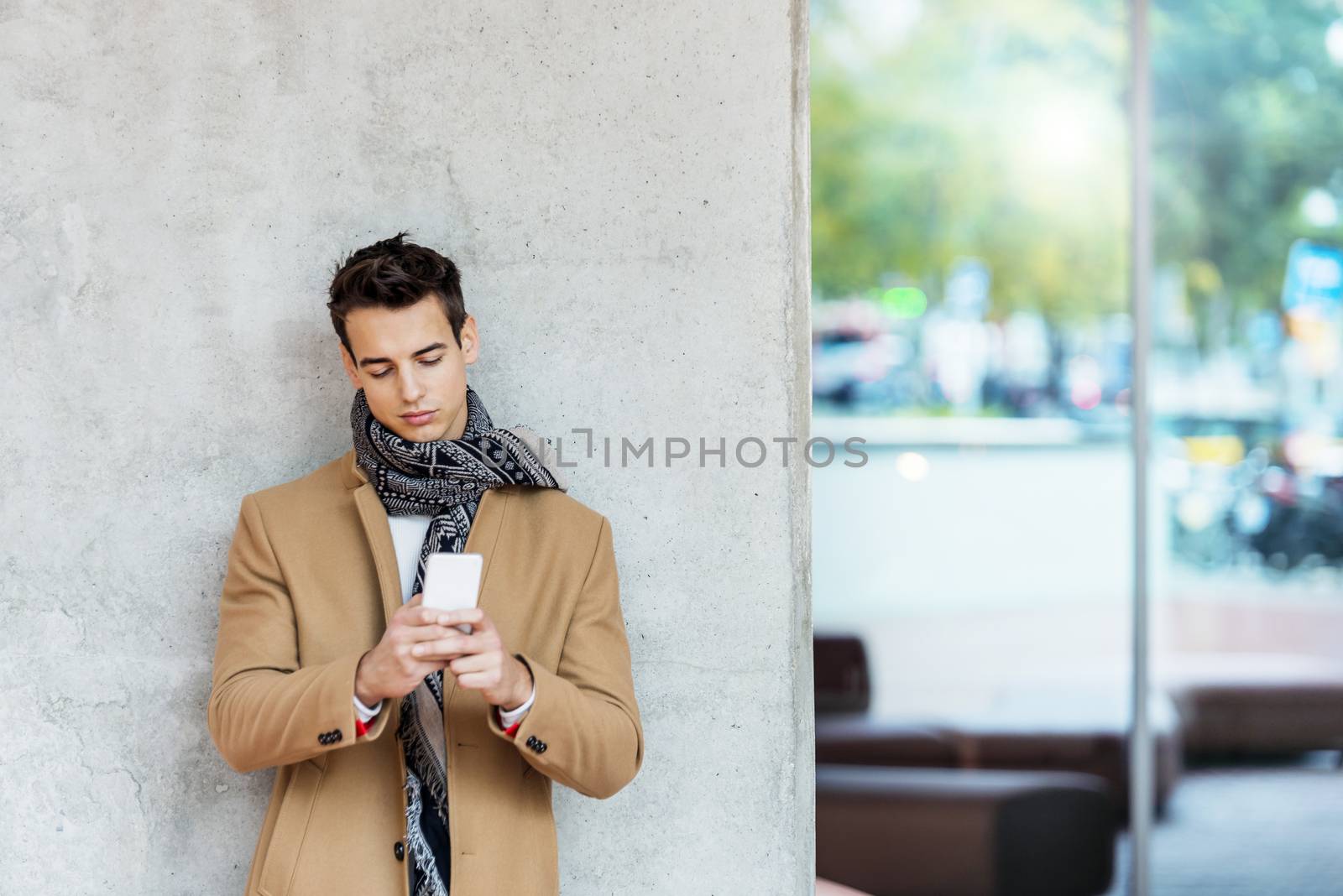 Front view of stylish young man wearing denim clothes leaning on a wall while using a mobile phone outdoors by raferto1973