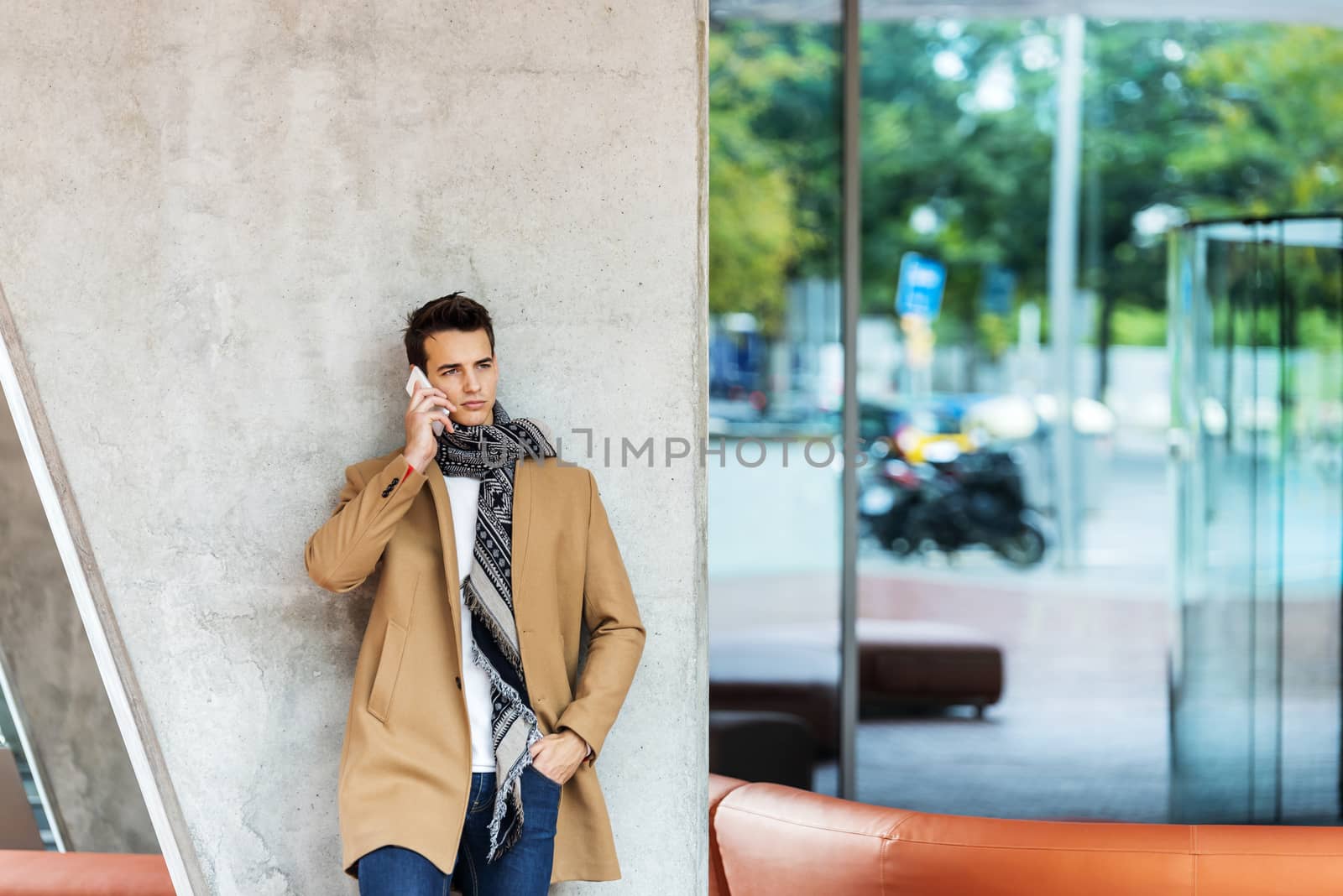 Front view of stylish young man wearing denim clothes leaning on a wall while using a mobile phone outdoors by raferto1973