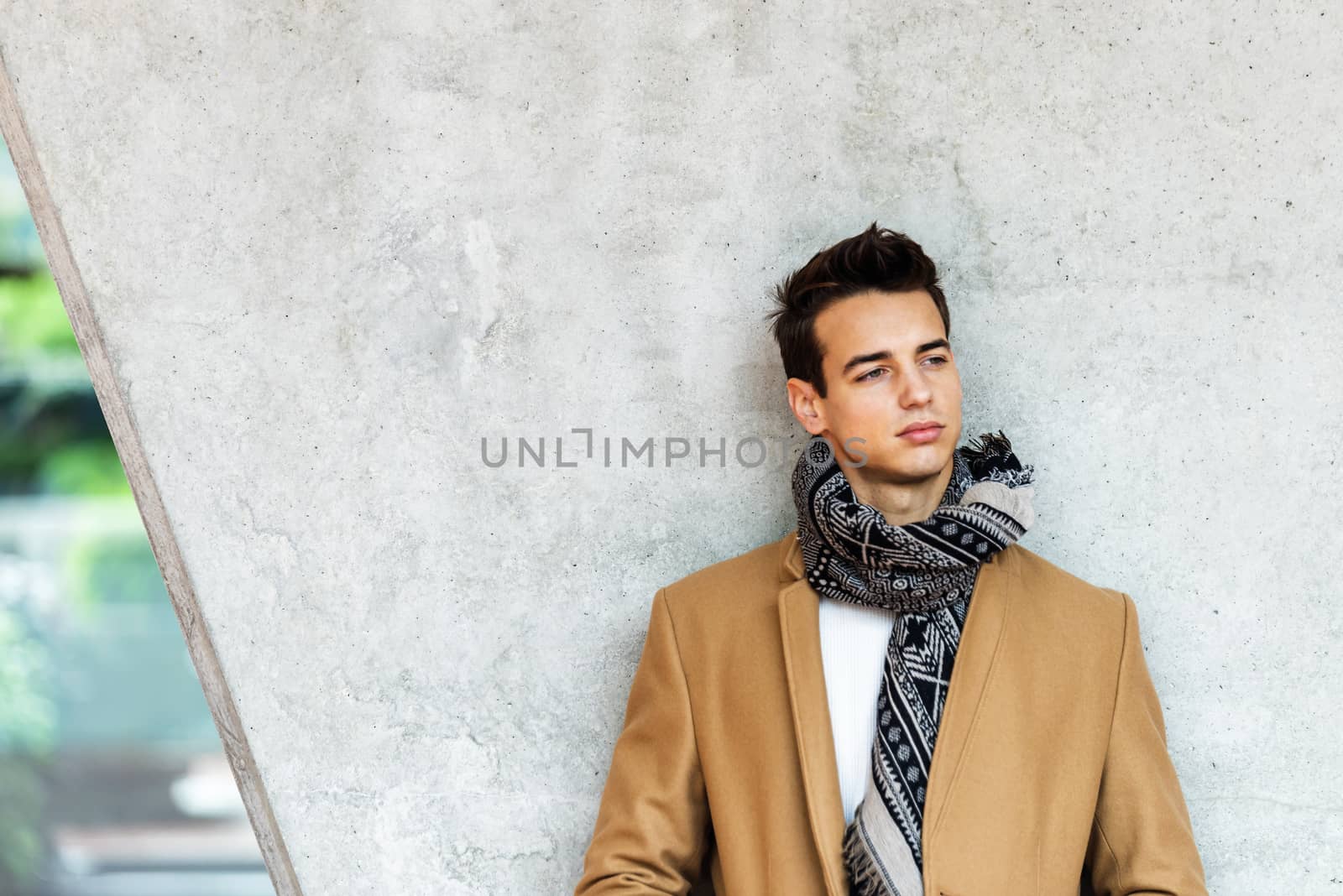 Front view of stylish young man wearing coat and scarf leaning on a wall while looking away