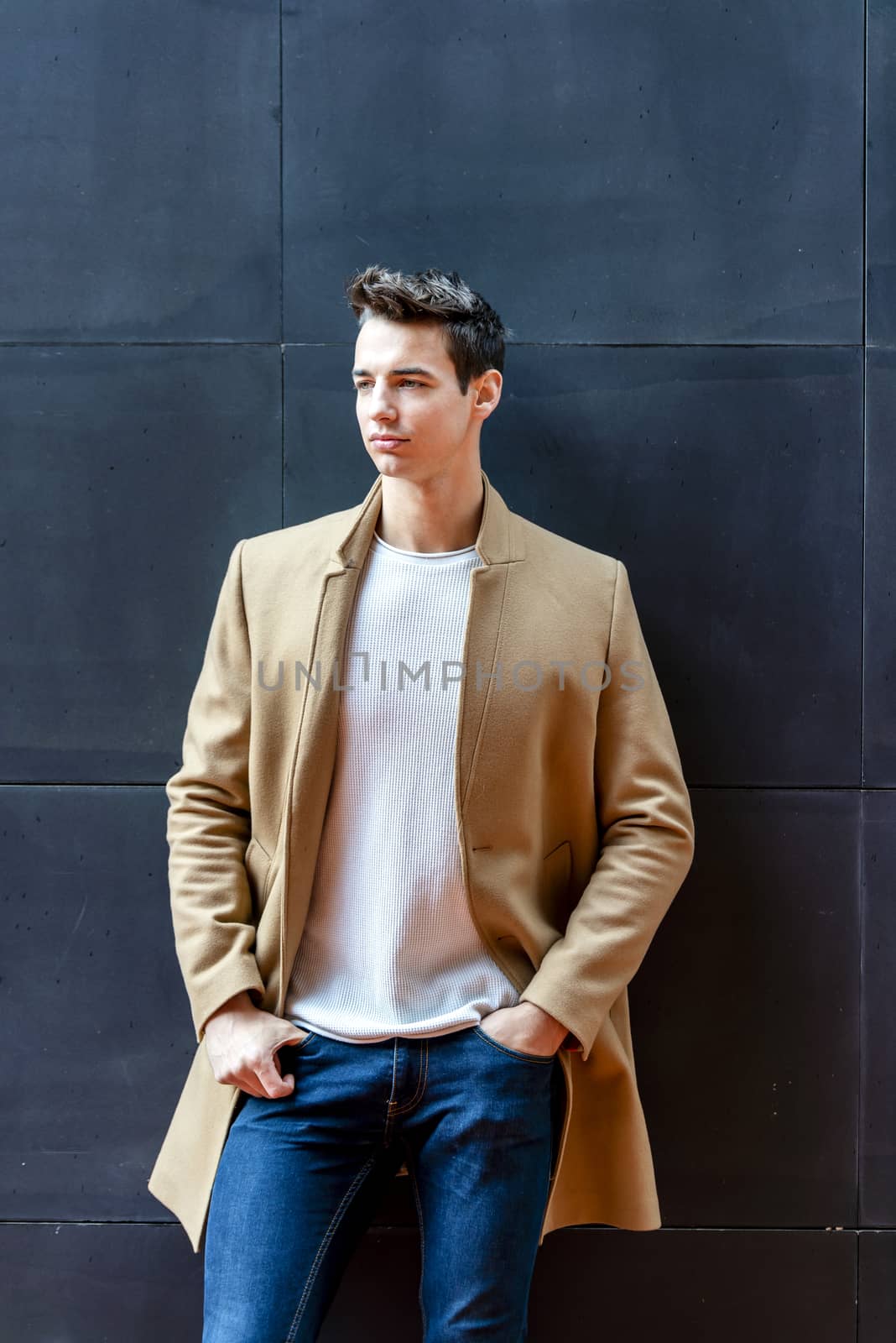 Front view of a trendy young man standing against black wall while looking away outdoors in the street