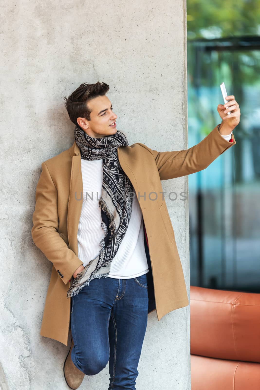 Side view of young trendy man leaning on a wall outdoors wearing denim clothes while taking a selfie in sunny day