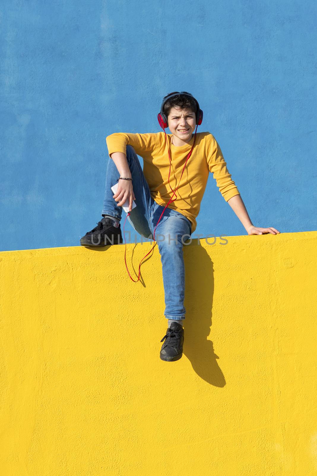 Front view of a young boy wearing casual clothes sitting on a yellow fence against a blue wall while using a mobile phone to listening music