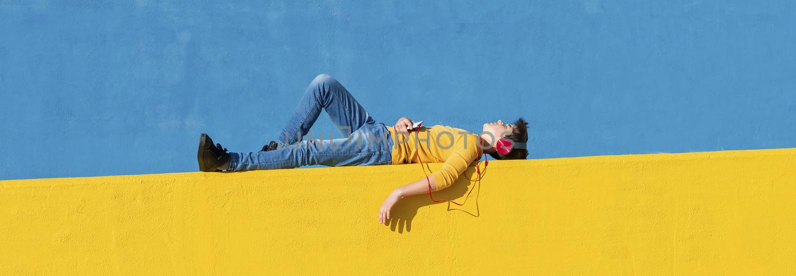 Front view of a young boy wearing casual clothes lying on a yellow fence against a blue wall while using a mobile phone to listening music by headphones by raferto1973