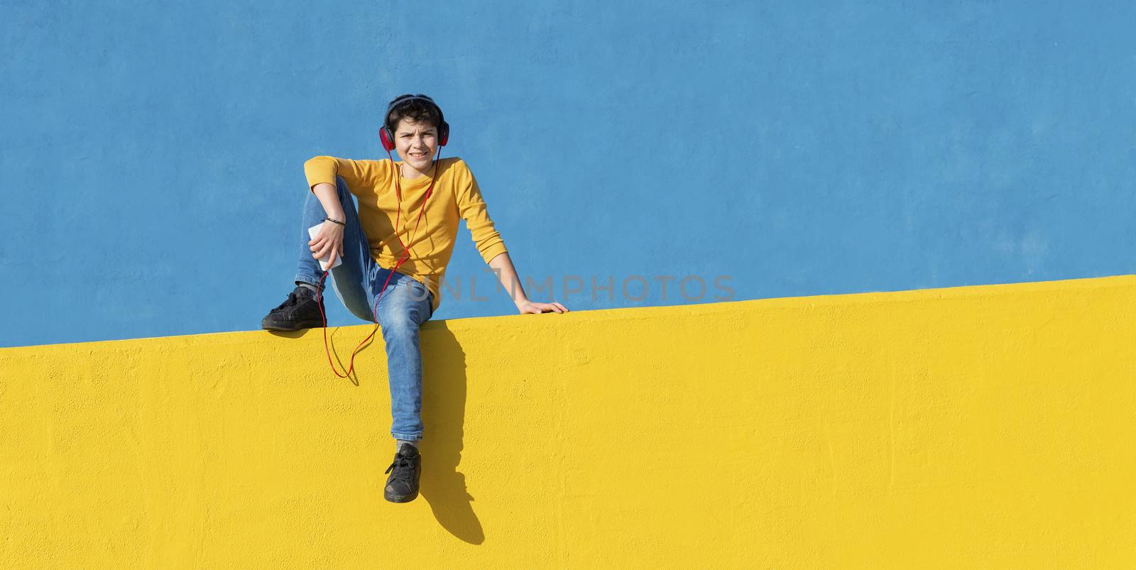 Front view of a young boy wearing casual clothes sitting on a yellow fence against a blue wall while using a mobile phone to listening music