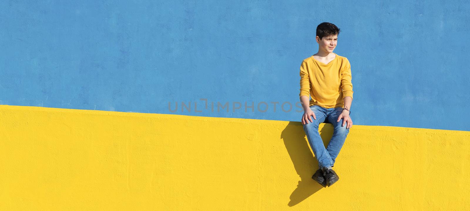 Front view of a young boy wearing casual clothes sitting on a yellow fence against a blue wall in a sunny day