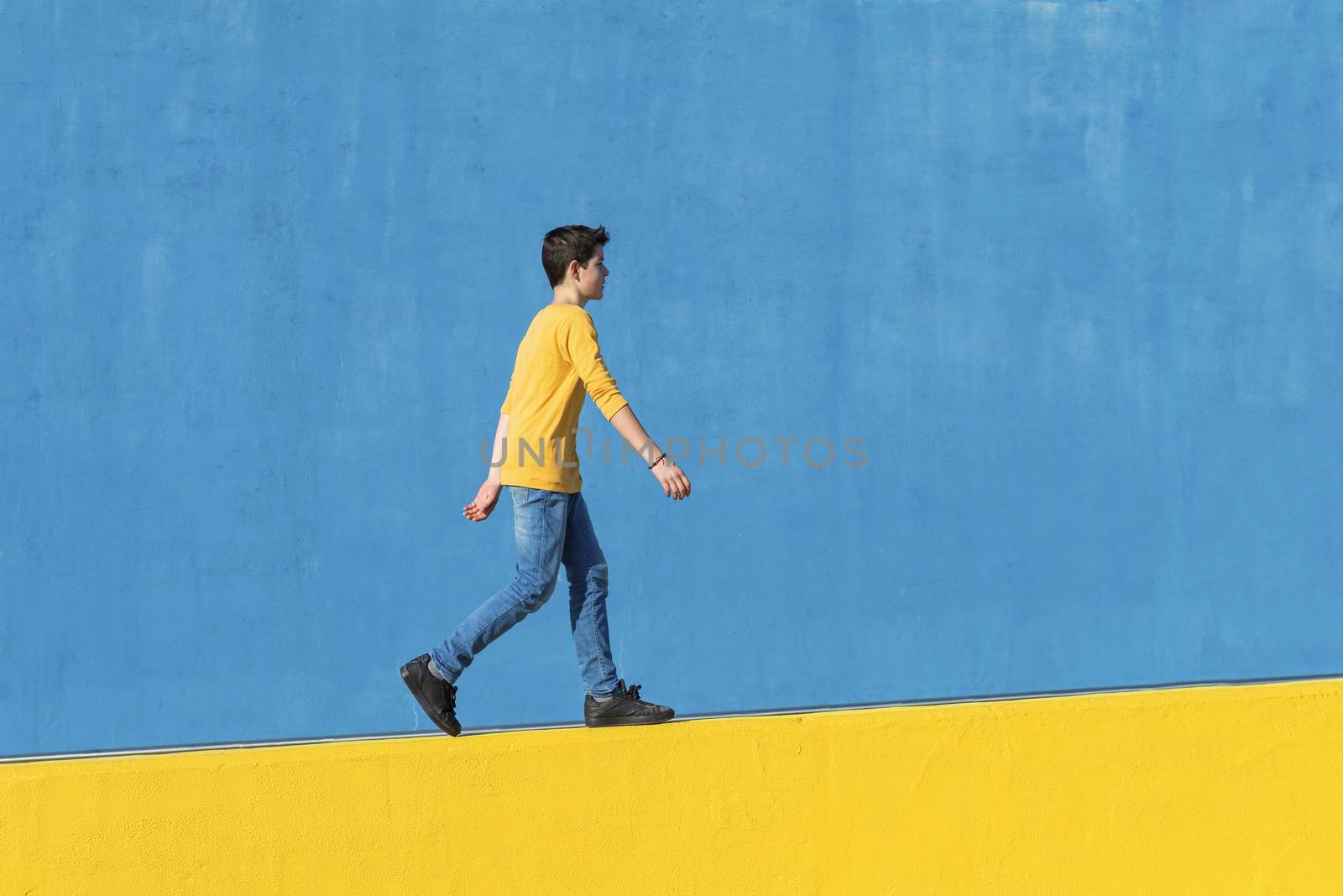 Young boy wearing casual clothes walking against a blue wall in a sunny day