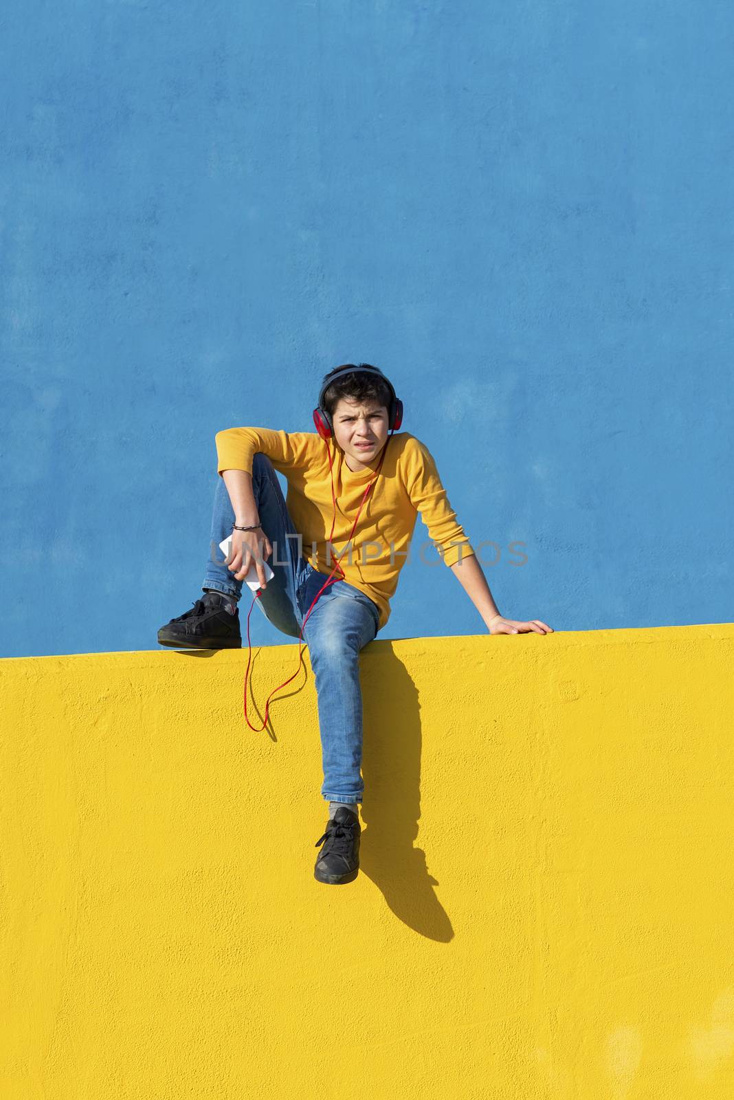 Front view of a young boy wearing casual clothes sitting on a yellow fence against a blue wall while using a mobile phone to listening music