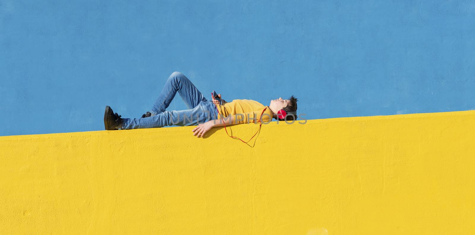 Front view of a young boy wearing casual clothes lying on a yellow fence against a blue wall while using a mobile phone to listening music by headphones by raferto1973