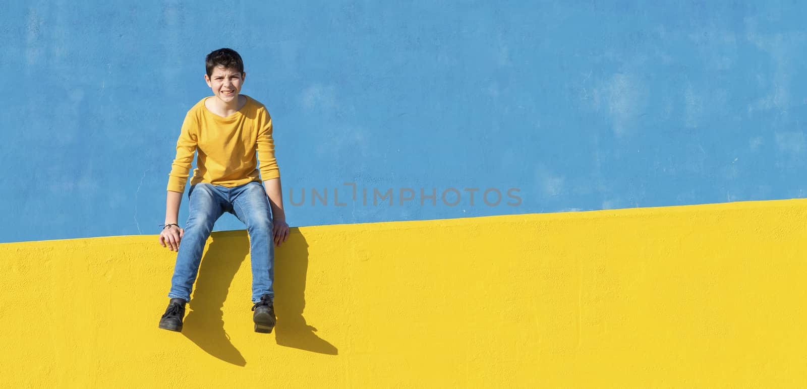 Front view of a young boy wearing casual clothes sitting on a yellow fence against a blue wall