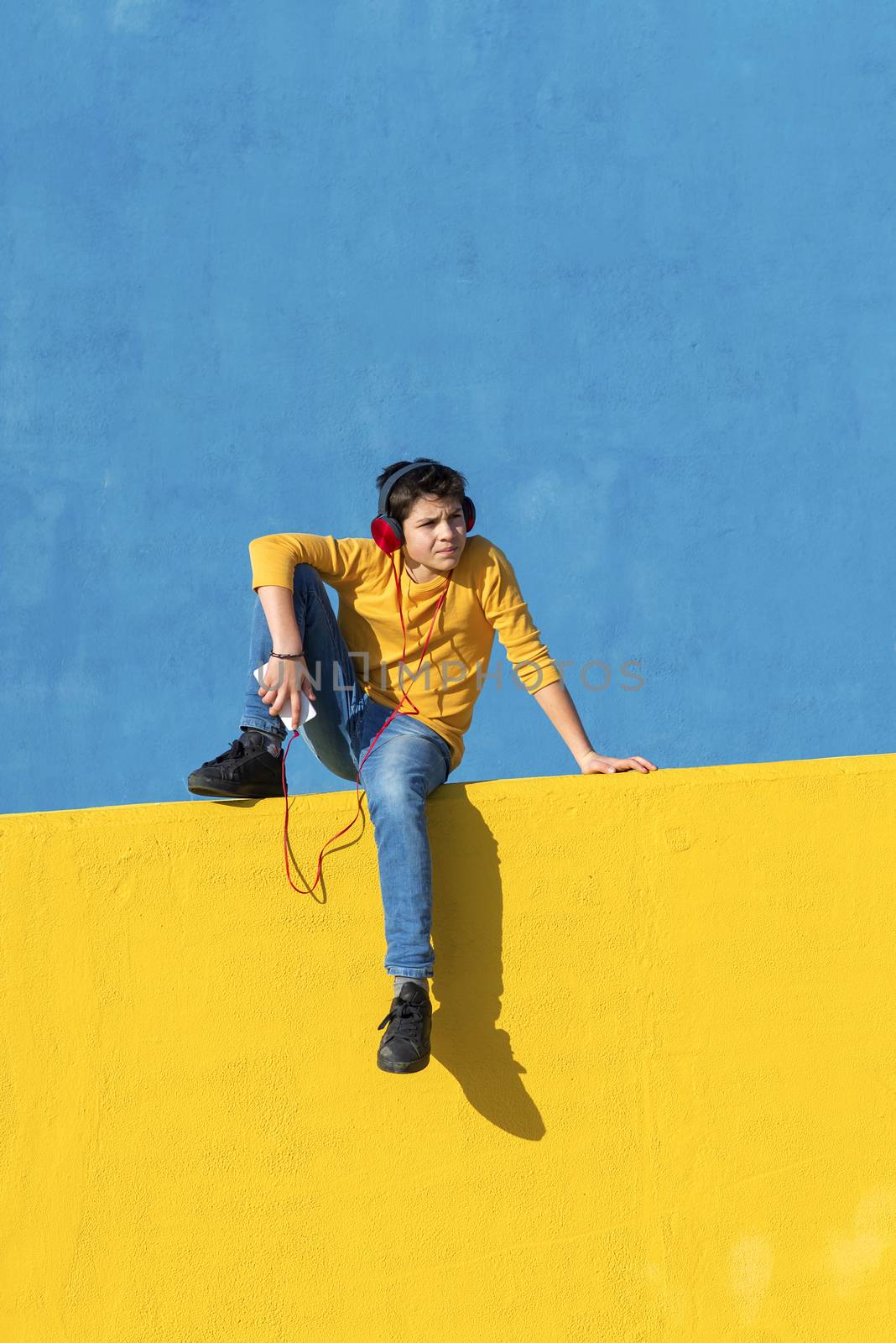 Front view of a young boy wearing casual clothes sitting on a yellow fence against a blue wall while using a mobile phone to listening music