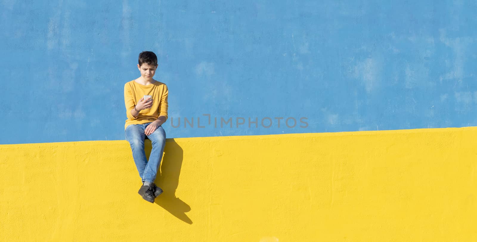 Front view of a young boy wearing casual clothes sitting on a yellow fence against a blue wall while using a mobile phone