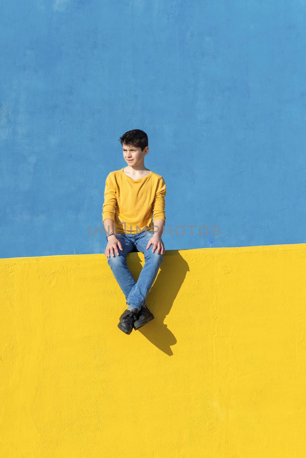 Front view of a young boy wearing casual clothes sitting on a yellow fence against a blue wall in a sunny day