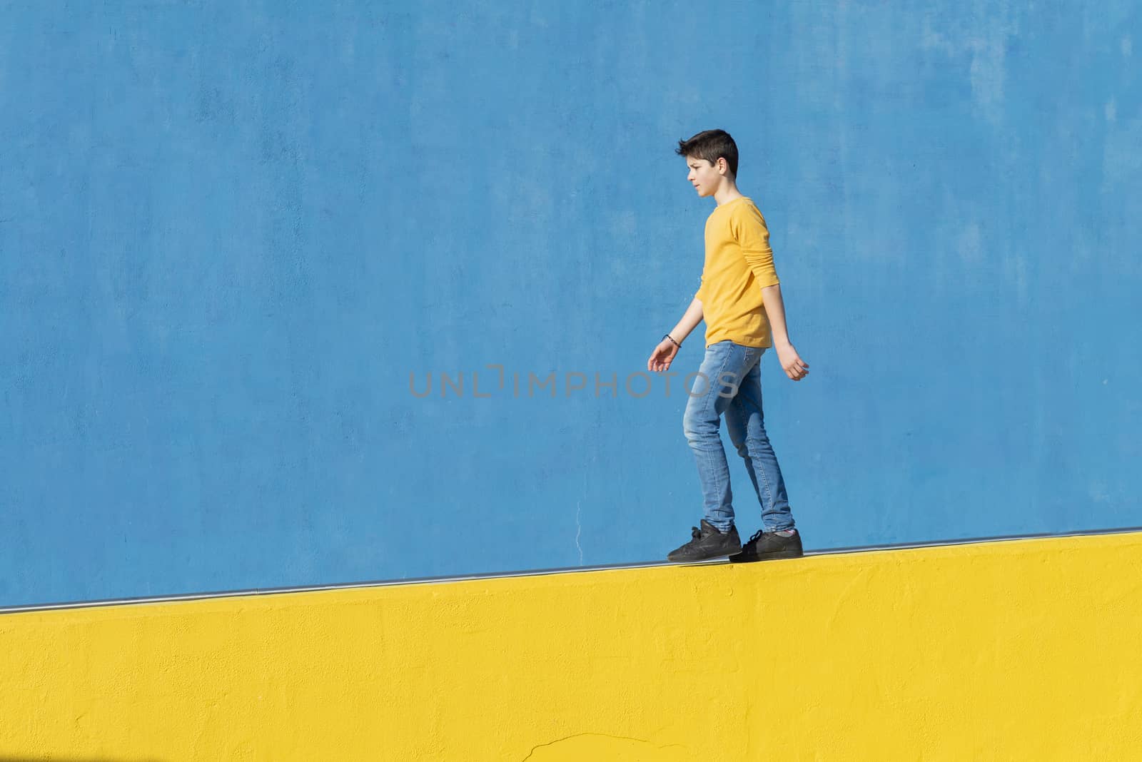 Young boy wearing casual clothes walking against a blue wall in a sunny day