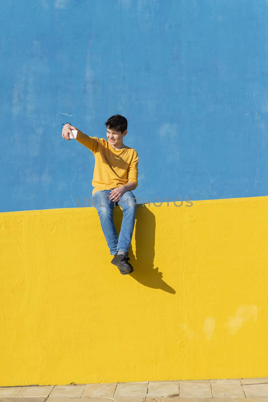 Front view of a young boy wearing casual clothes sitting on a yellow fence against a blue wall while using a mobile phone