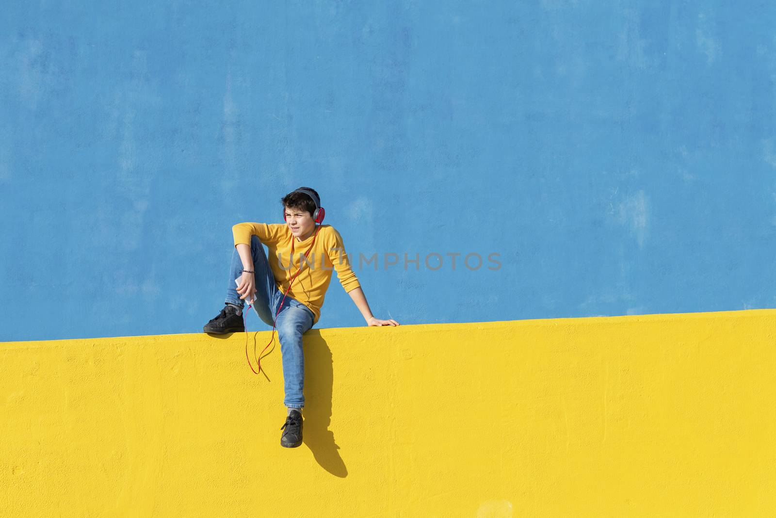 Front view of a young boy wearing casual clothes sitting on a yellow fence against a blue wall while using a mobile phone to listening music