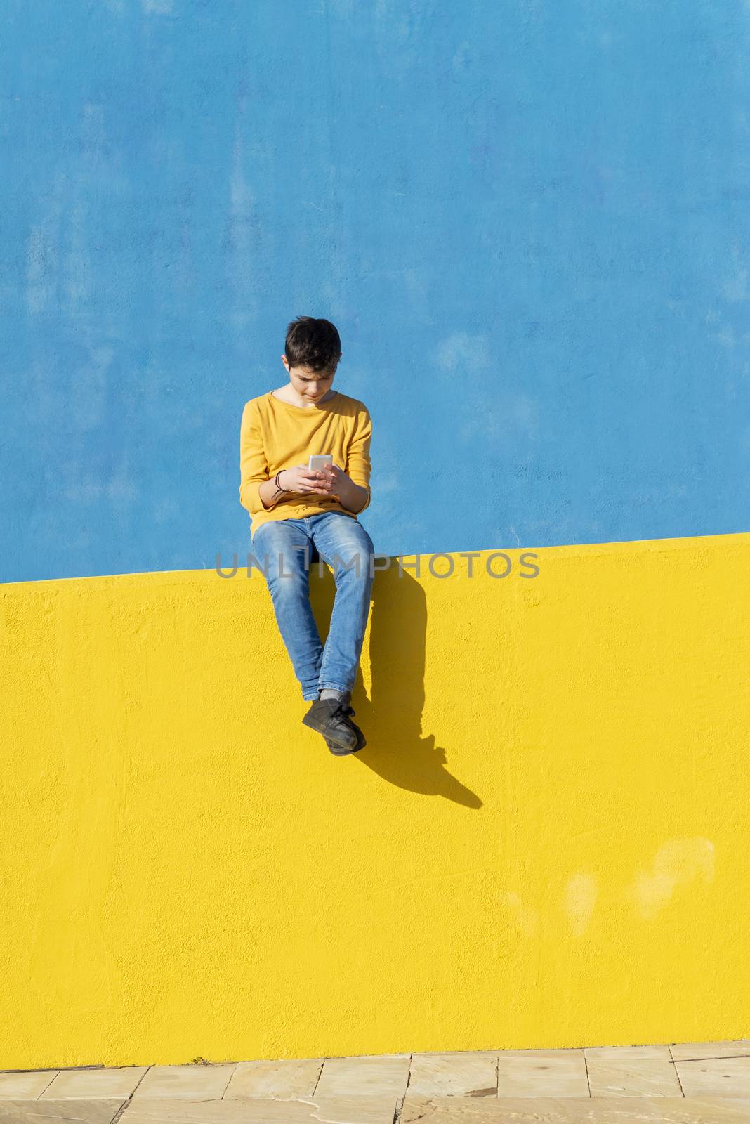 Front view of a young boy wearing casual clothes sitting on a yellow fence against a blue wall while using a smartphone by raferto1973