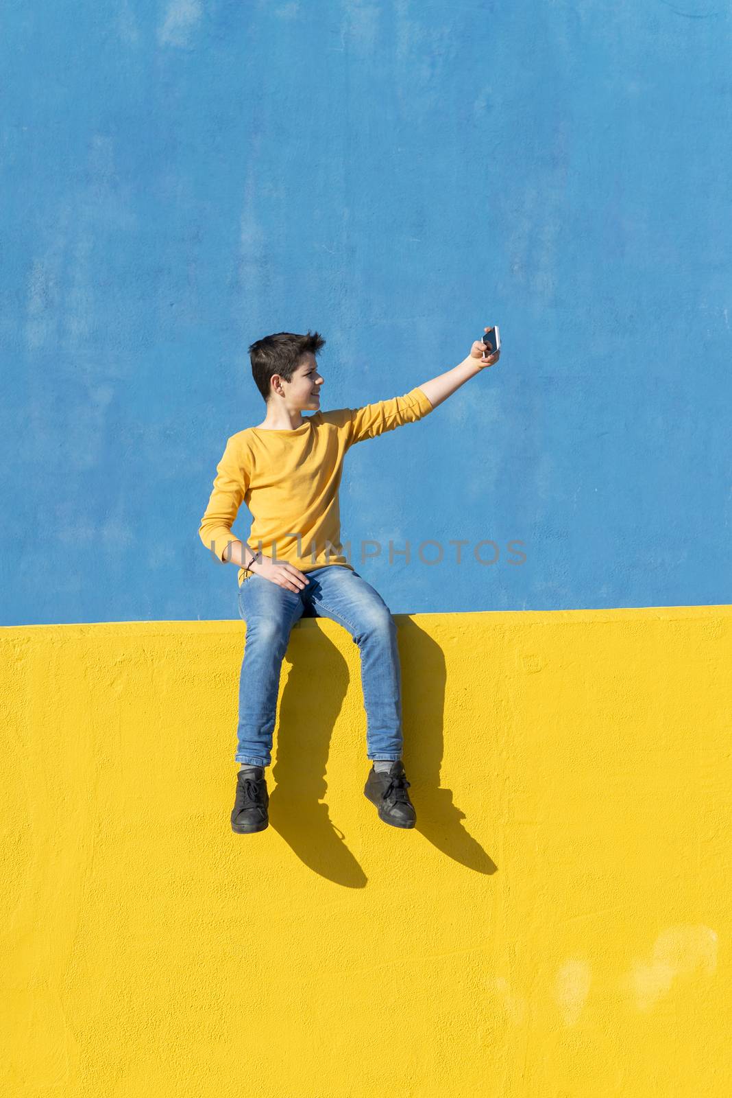 Front view of a young boy wearing casual clothes sitting on a yellow fence against a blue wall while using a mobile phone