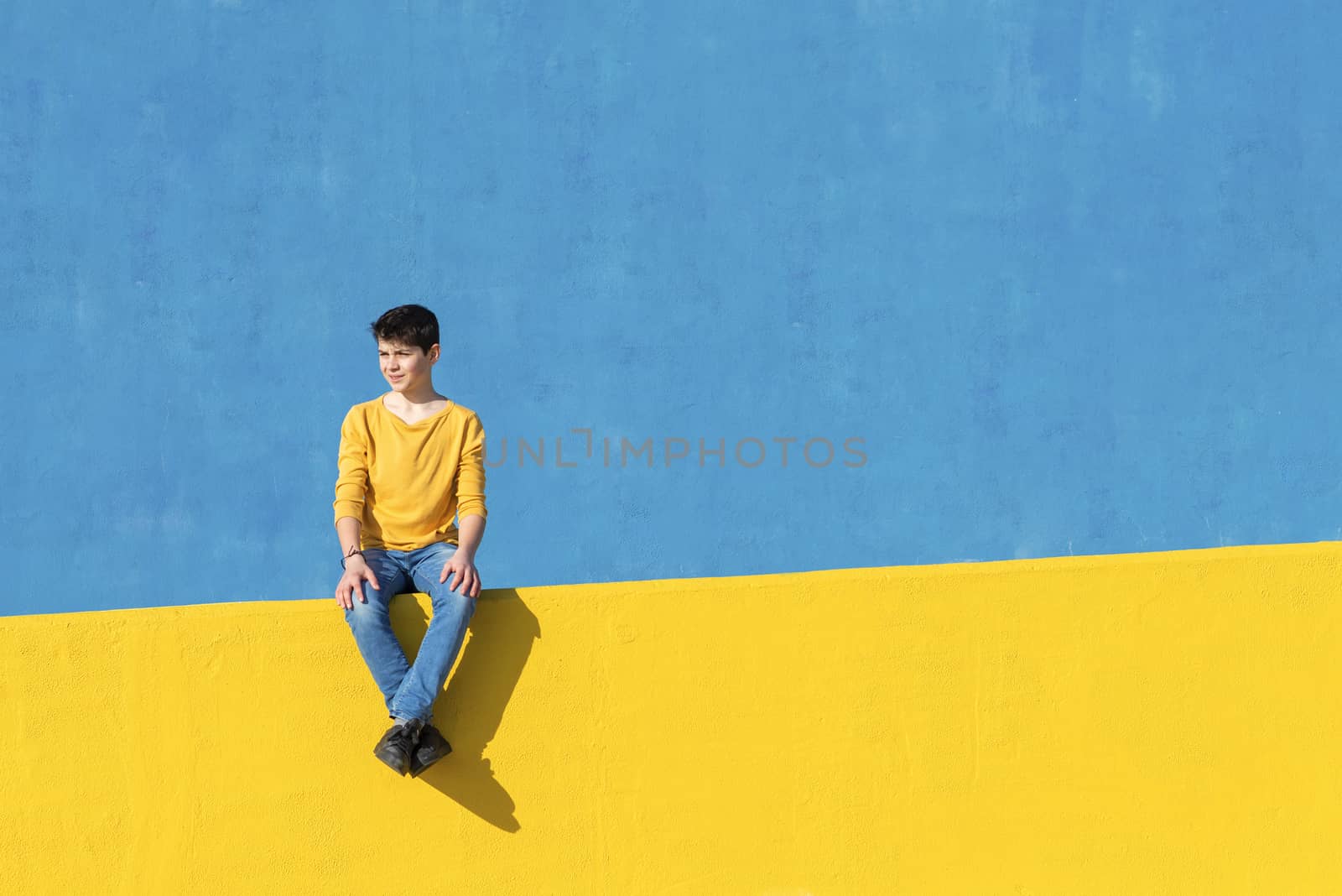 Front view of a young boy wearing casual clothes sitting on a yellow fence against a blue wall in a sunny day