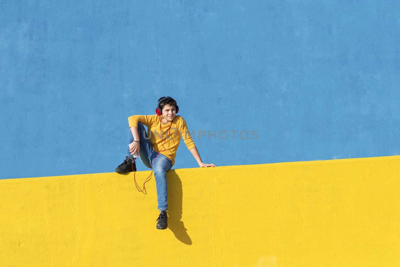 Front view of a young boy wearing casual clothes sitting on a yellow fence against a blue wall while using a mobile phone to listening music