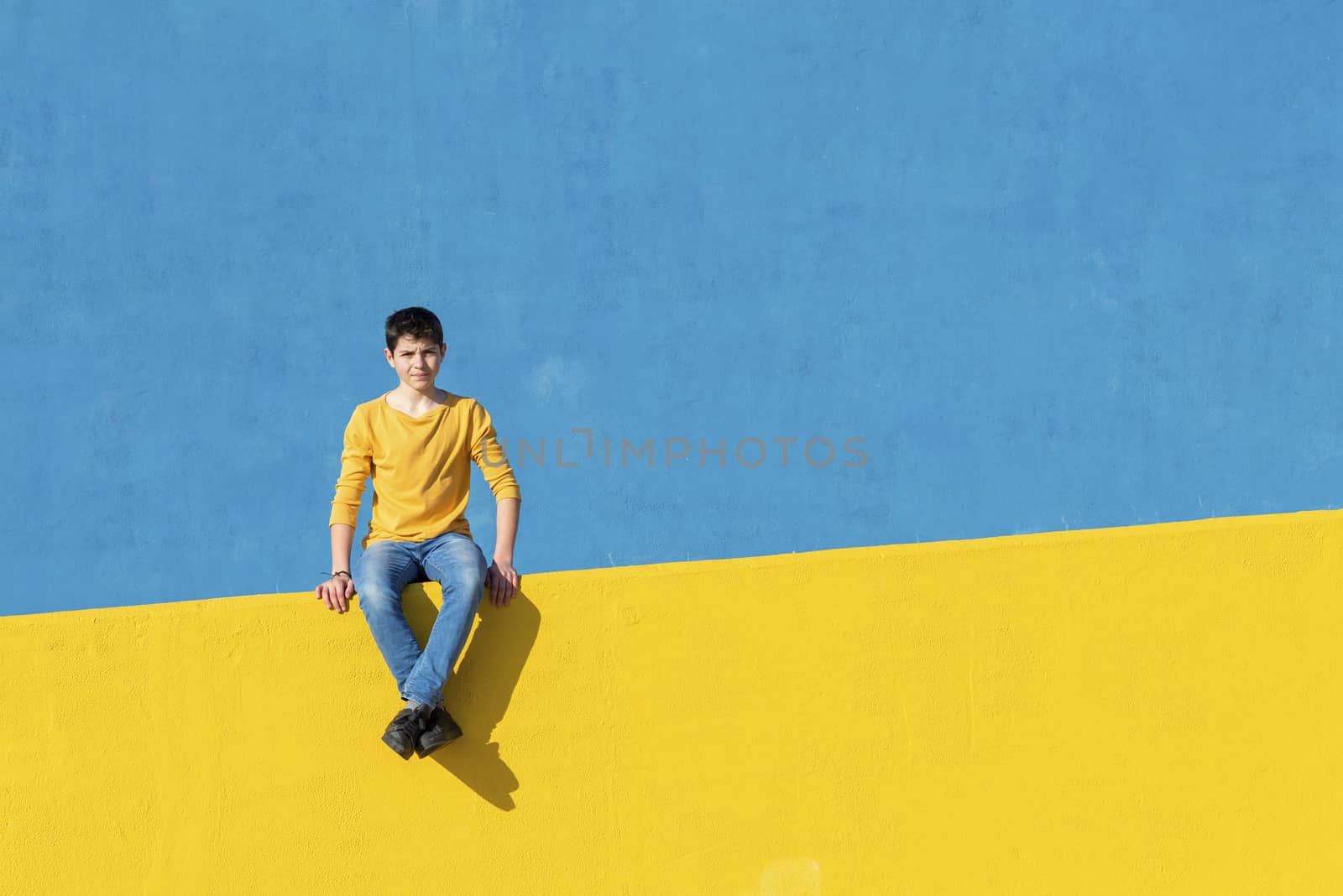 Front view of a young boy wearing casual clothes sitting on a yellow fence against a blue wall in a sunny day by raferto1973