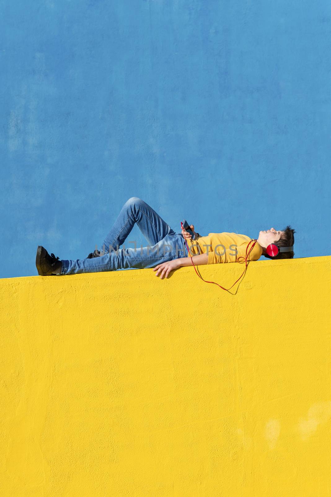 Front view of a young boy wearing casual clothes lying on a yellow fence against a blue wall while using a mobile phone to listening music