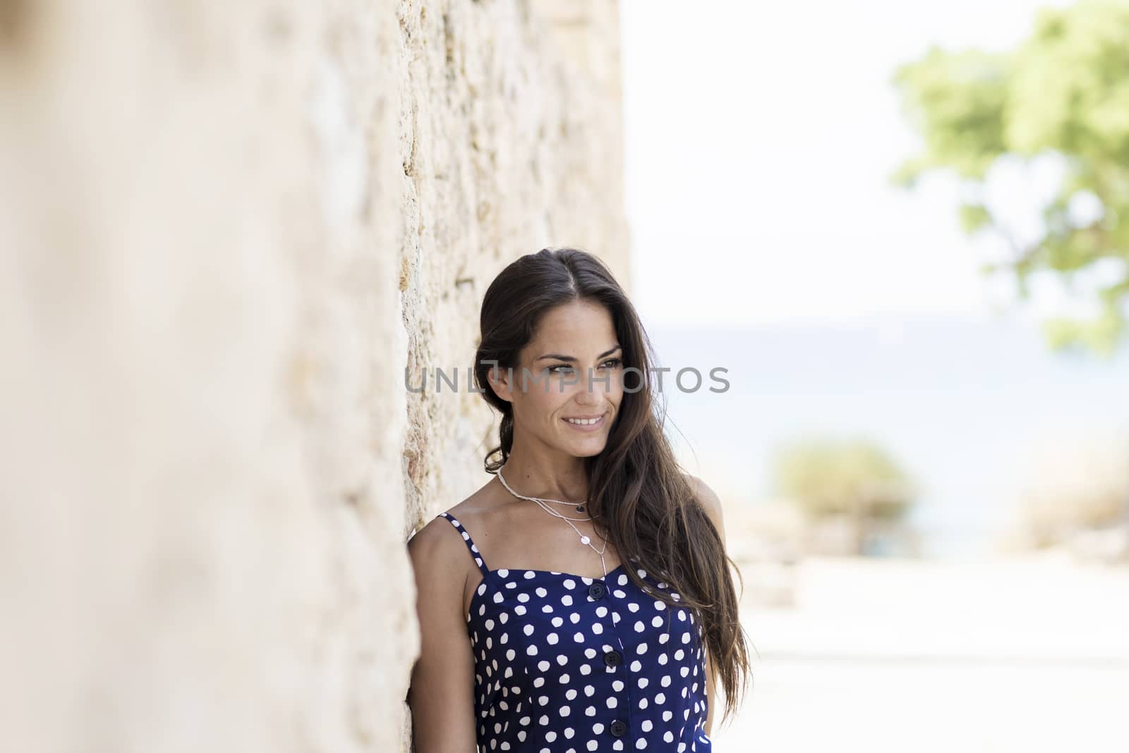 Beautiful smiling woman in blue dress leaning on wall while looking away