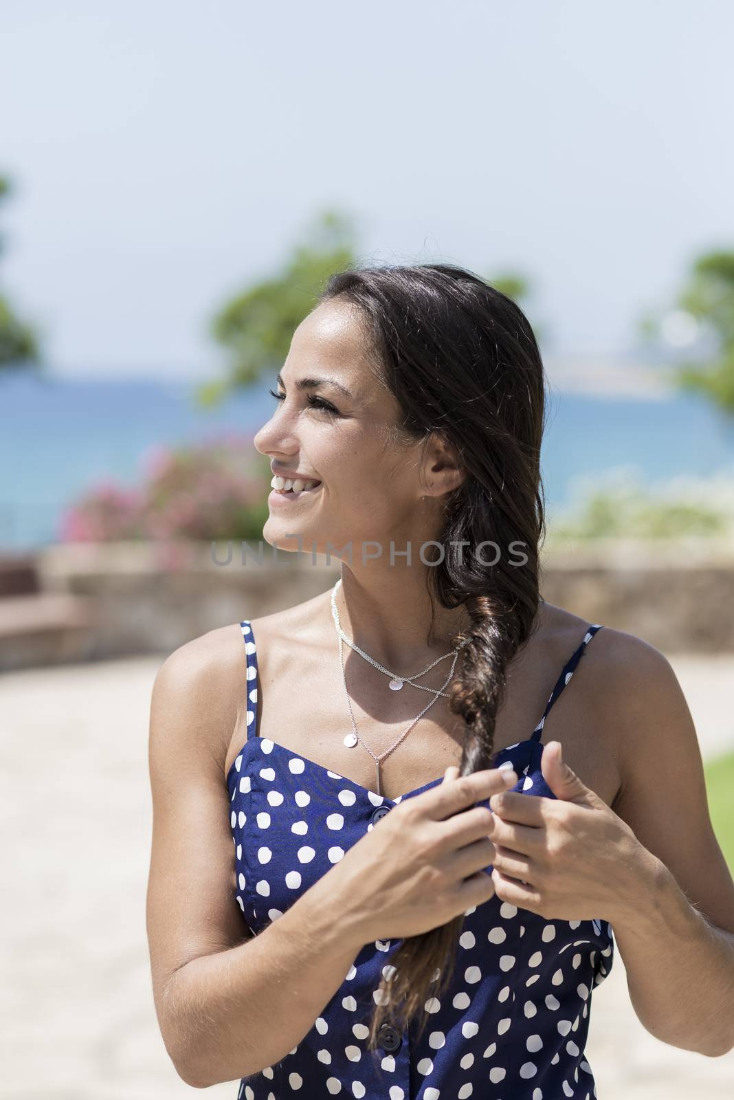 Beautiful smiling woman in blue dress standing against sea while touching hair