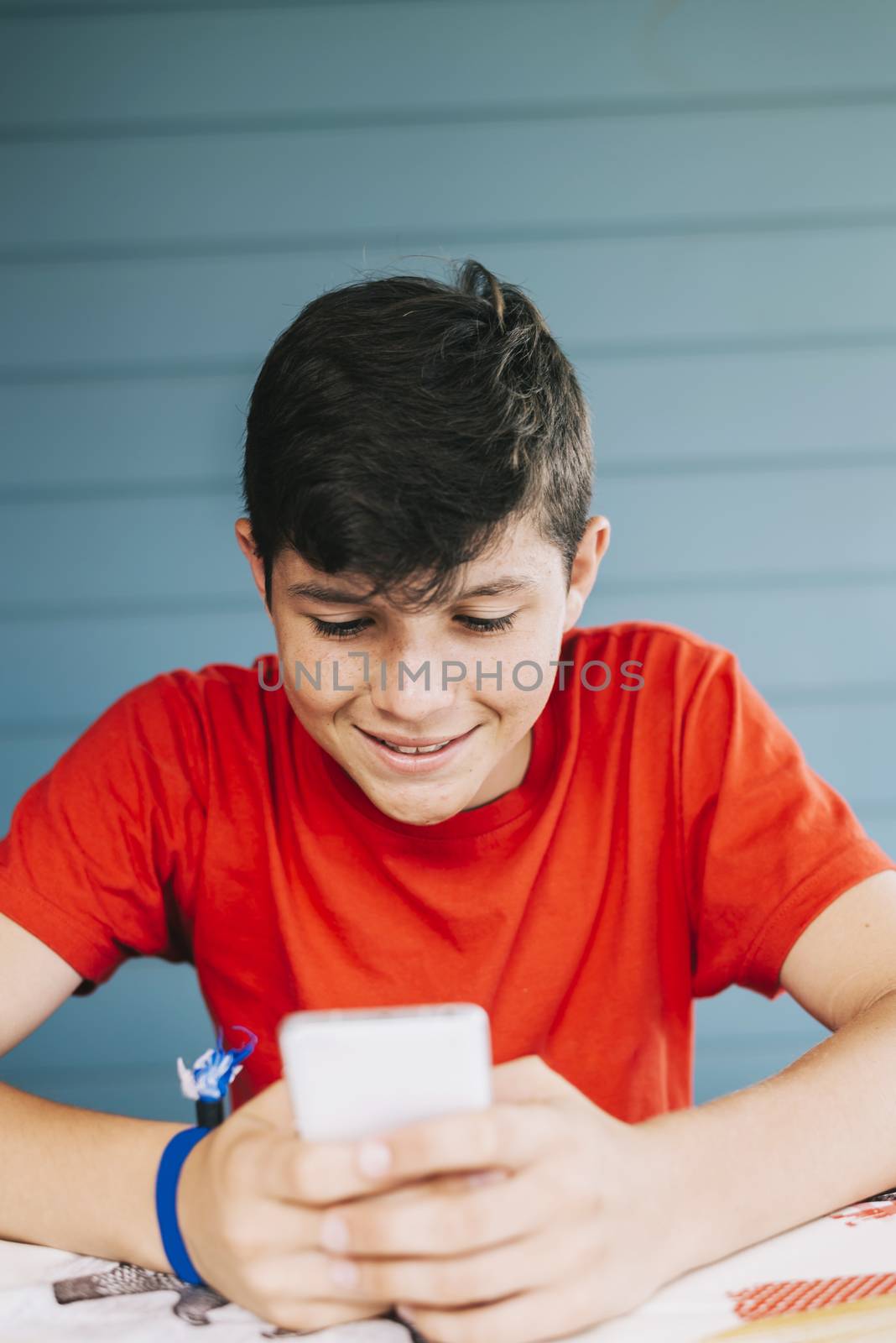 Handsome Caucasian 13-year old boy wearing red t-shirt sitting outdoors using electronic gadget. Schoolboy holding mobile phone, messaging friends