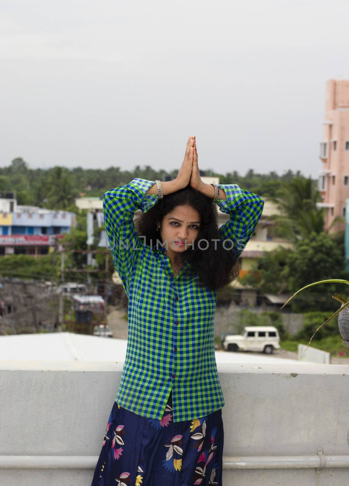 A young Hindu woman standing in an open environment, eyes opened and praying with her hands above the head to the Sun God by 9500102400