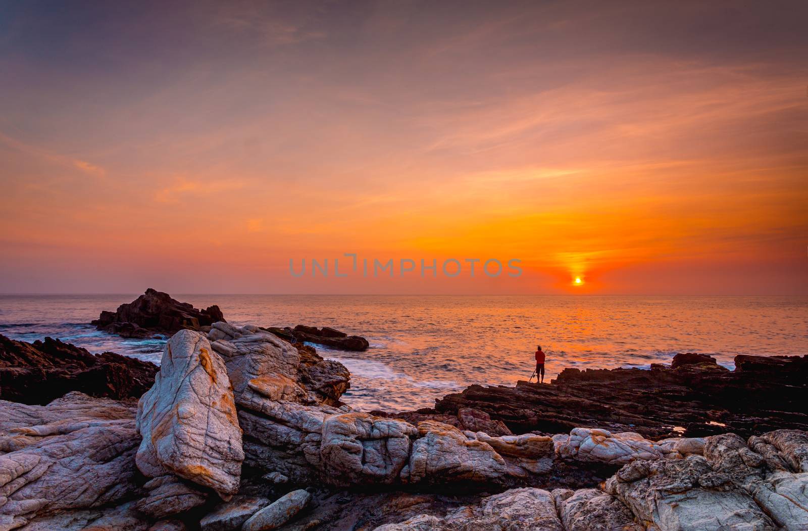 Brilliant vivid orange sunrise over the ocean with foreground rocks in various textures and stained with orange ochres.  A fisherman in the distant rocks hopes for a morning catch
