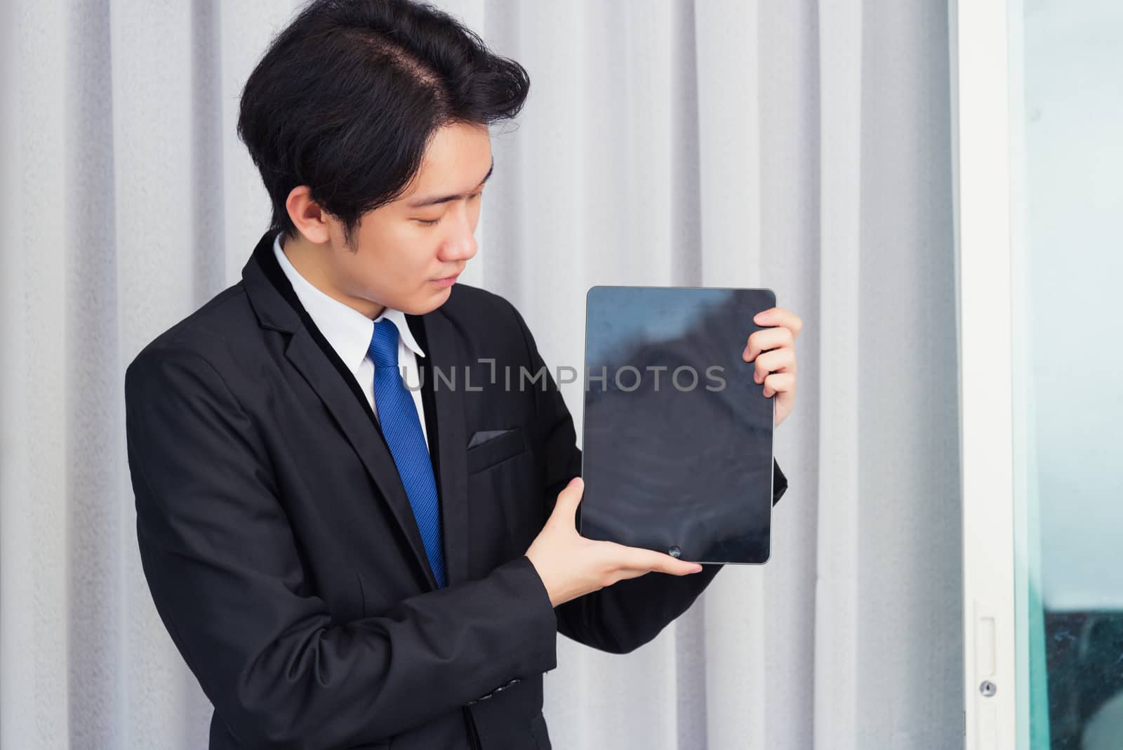 Work from home, Asian young businessman wear suit video conference call or facetime he smiling and showing blcnk screen of smart digital tablet computer at home office, looking to tablet