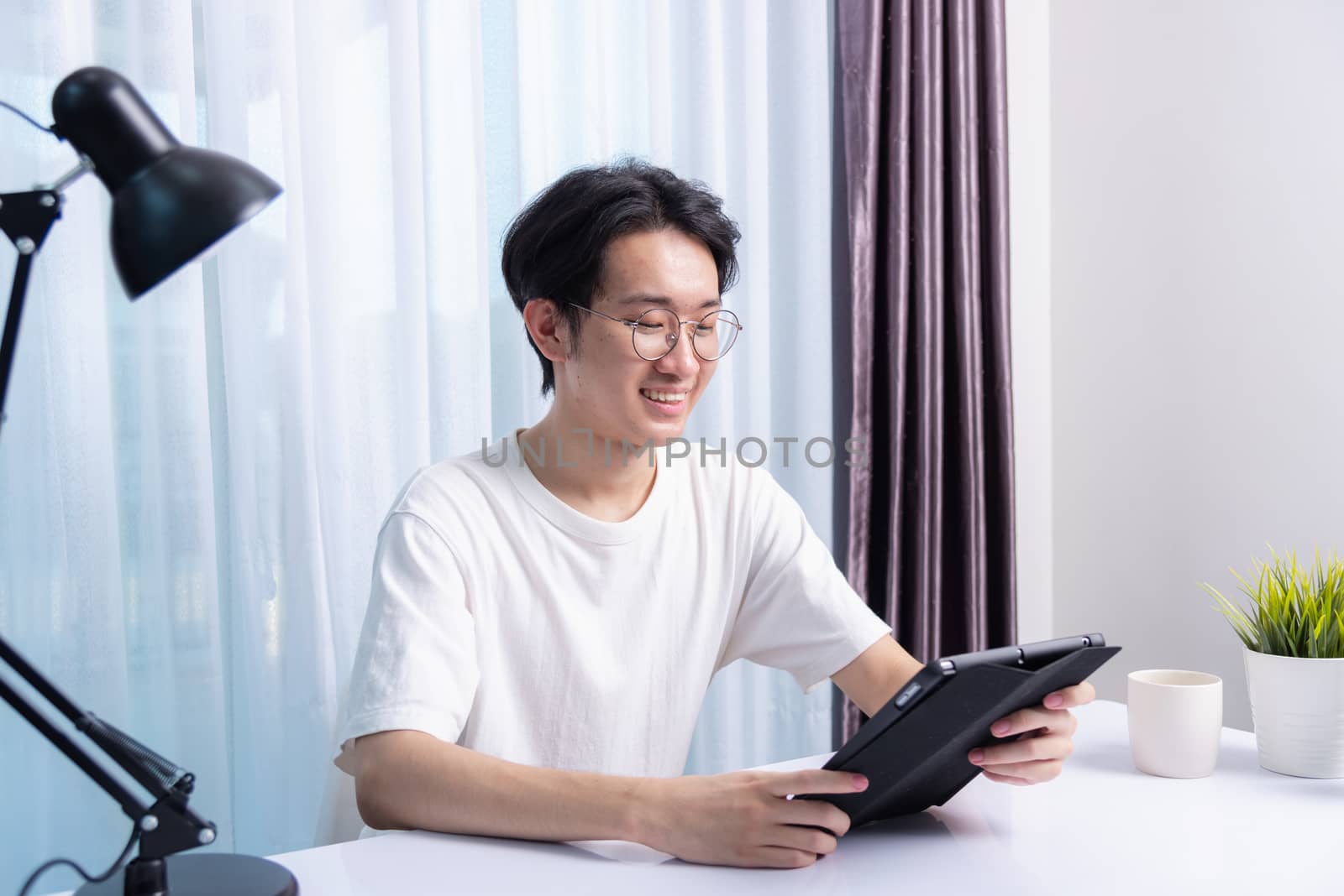 Happy Asian young business handsome man work from home office wear glasses, t-shirt comfortable he smiling and using a black modern smart digital tablet computer to read email or learning on desk