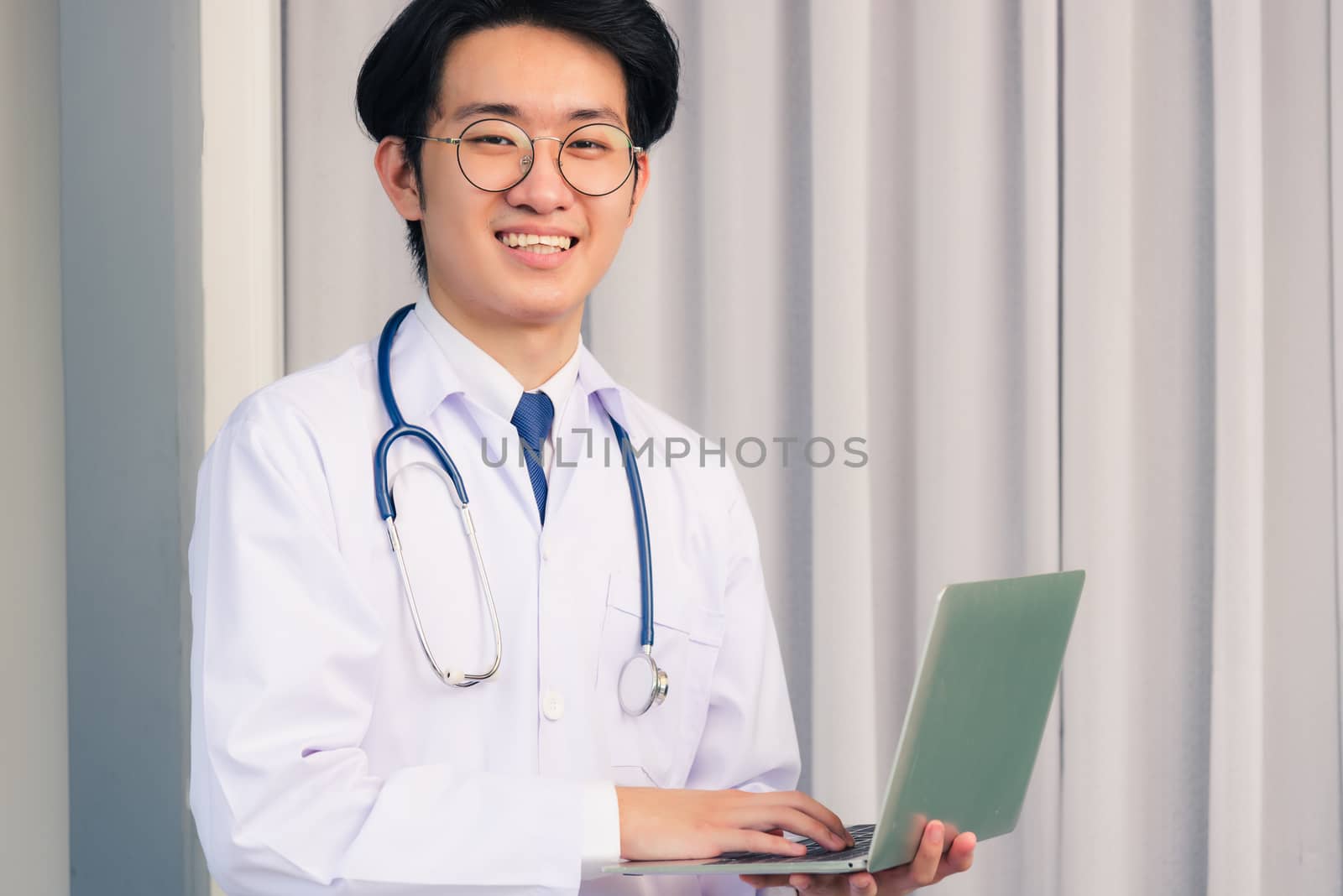 Portrait of Asian young handsome doctor man wearing a doctor's dress and stethoscope smiling sitting at hospital office, Health medical care concept