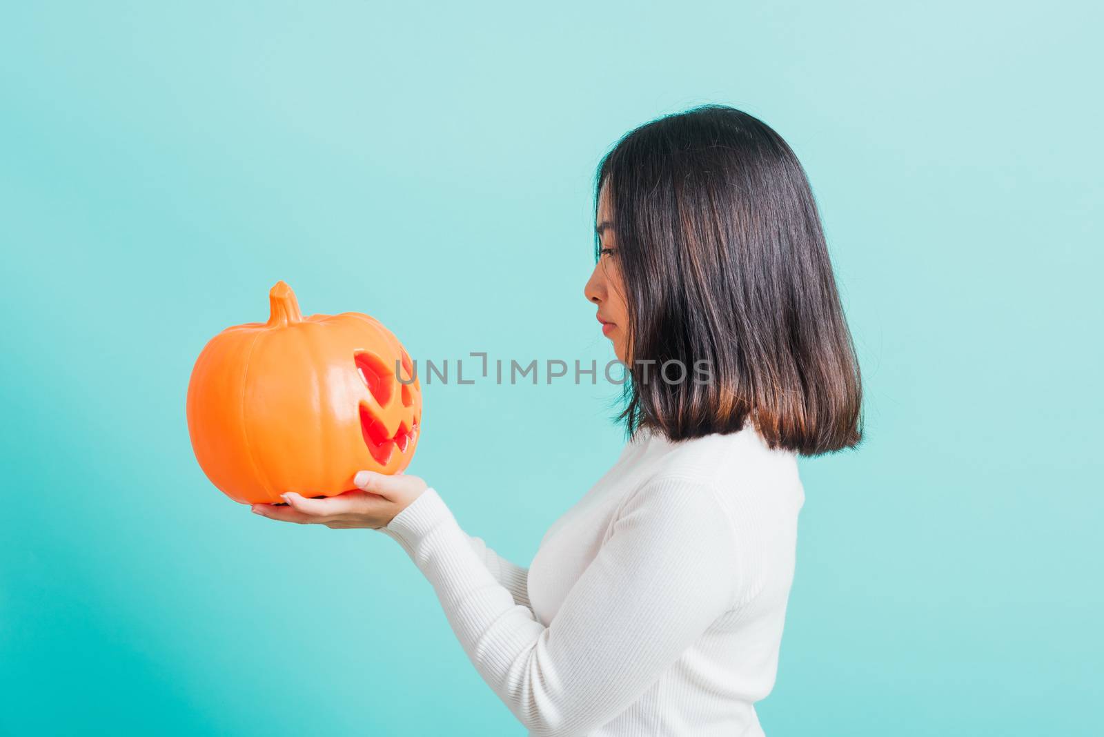 Portrait of Asian beautiful young woman holding orange model pumpkins and looking it, funny happy female with ghost pumpkins, studio shot isolated on blue background