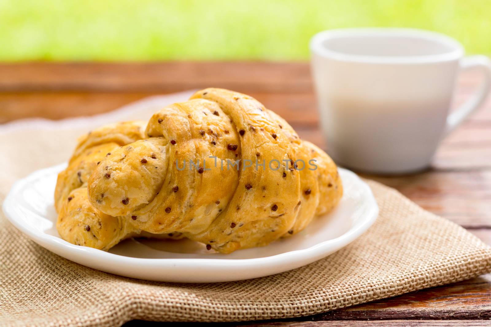 Breakfast concept. Croissants with perilla seeds on white plate and white cup of black coffee on wooden table with green bokeh background.