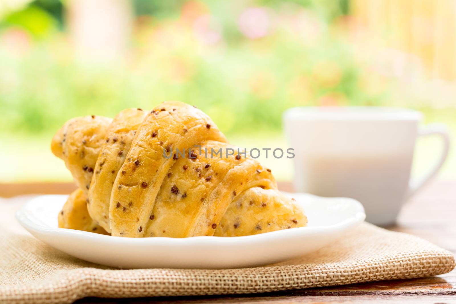 Breakfast concept. Croissants with perilla seeds on white plate and white cup of black coffee on wooden table with green bokeh background.