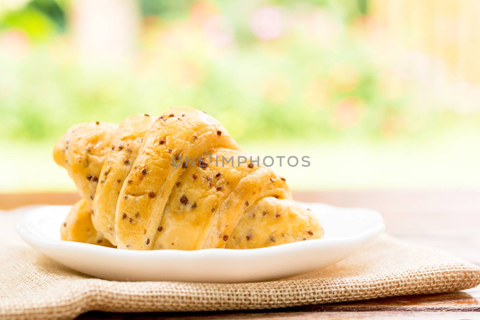 Breakfast concept. Croissants with perilla seeds on white plate and white cup of black coffee on wooden table with green bokeh background.