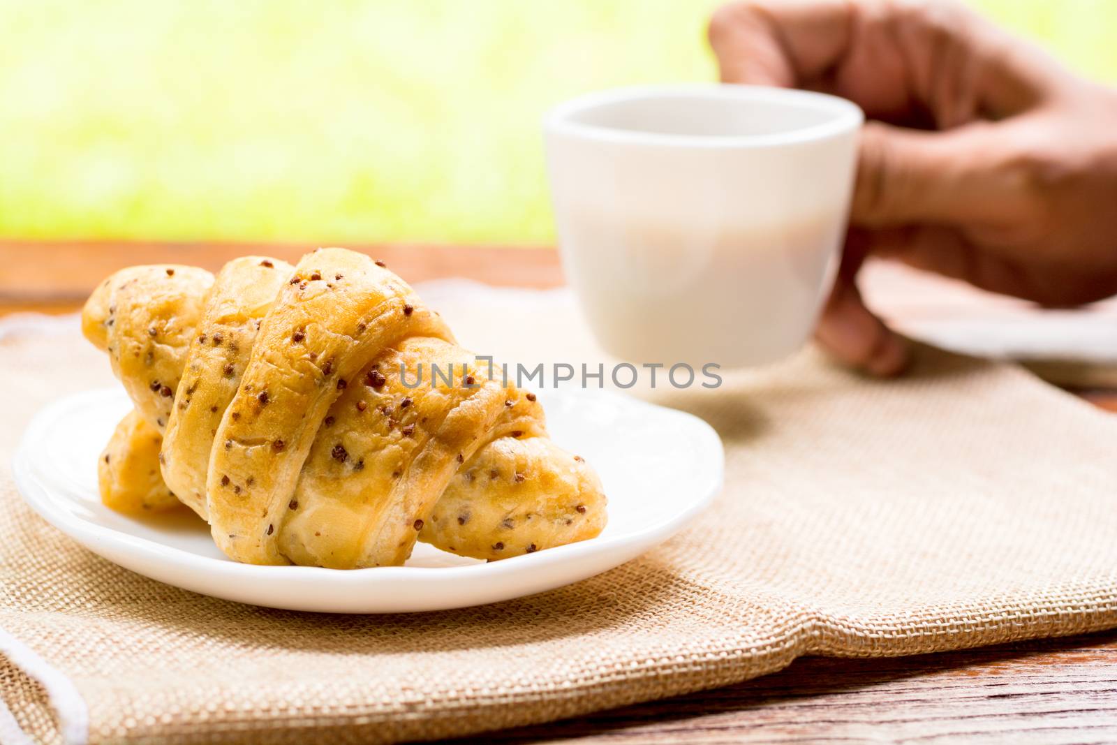 Breakfast concept. Croissants with perilla seeds on white plate and white cup of black coffee on wooden table with green bokeh background.