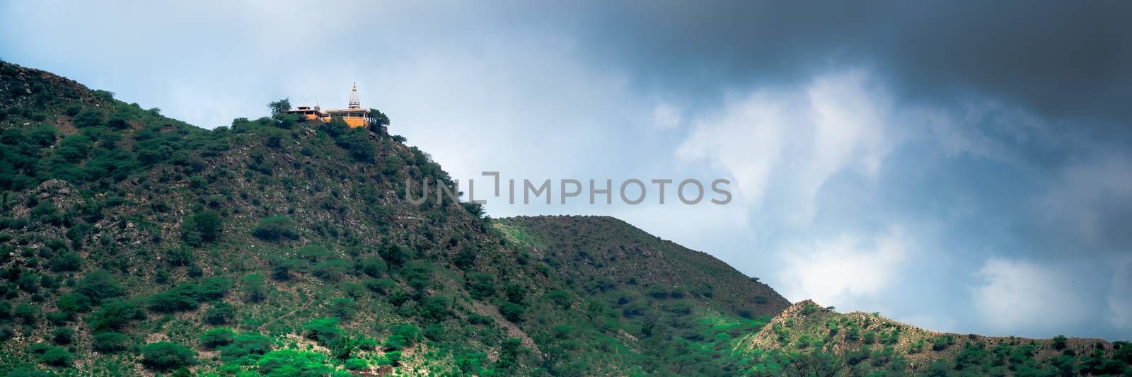 hindu temple on a hill covered in vegetation with the tower and spire clearly visible in the distance with a background of dark monsoon clouds shot in Rajasthan India. Shows the beautiful landmark locations used in the past to build temples and the amazing views from them